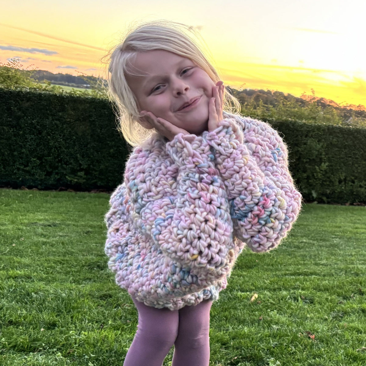 Barn i oversized hæklet Yarnicorn Sweater set forfra med armene løftet, så sweaterens rummelige ærmer og farverige masker ses.