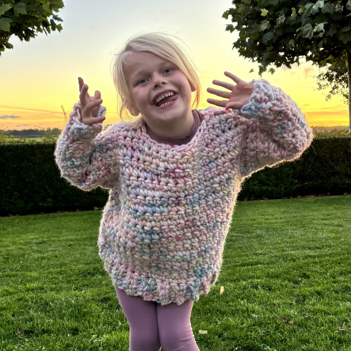 Barn i farverig, chunky hæklet børnesweater set fra siden med hånden i håret. Sweateren har lange ærmer og ribkant nederst.
