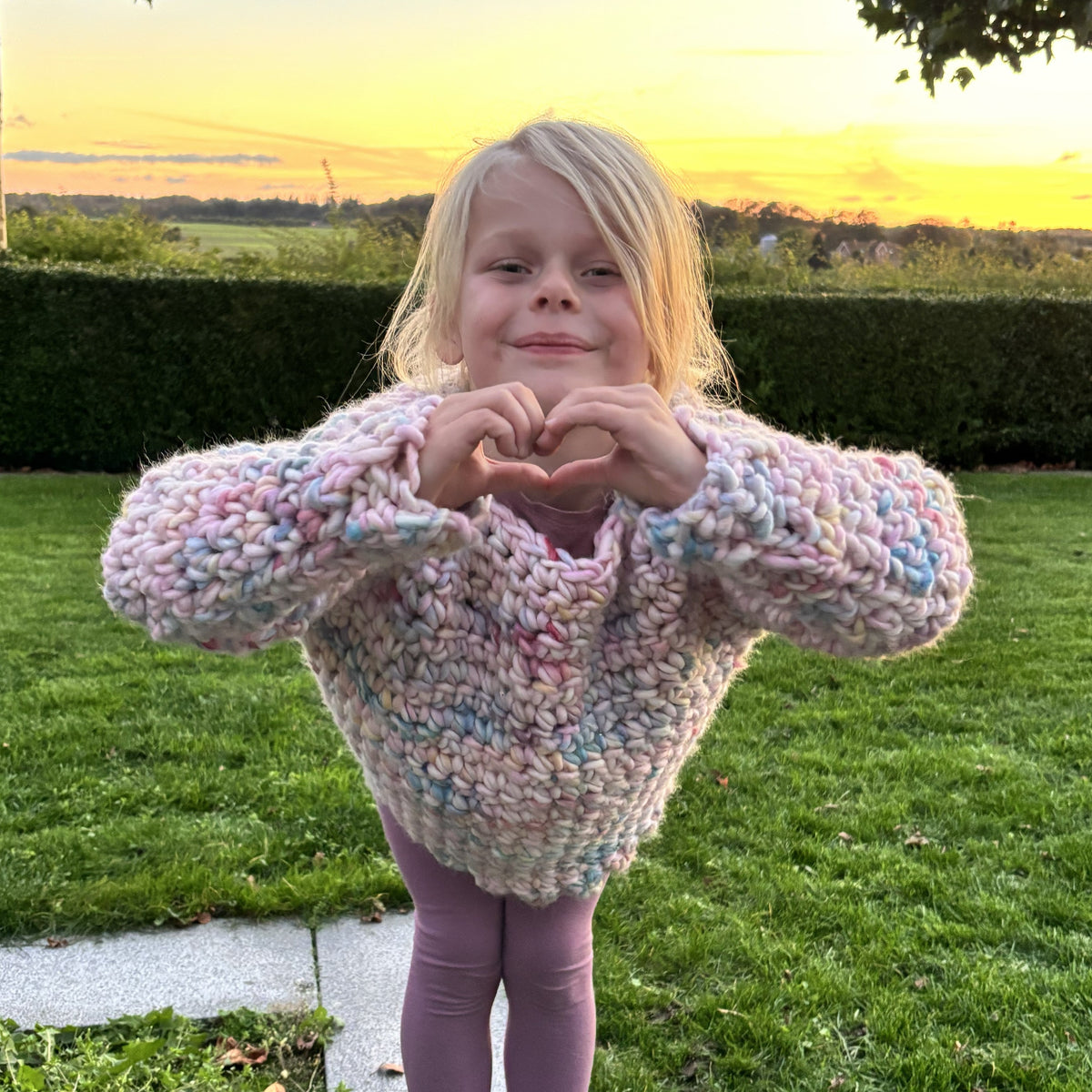 Pige fotograferet forfra i en farverig hæklet sweater med tykt garn, lange ærmer og hænderne samlet i et hjerte.