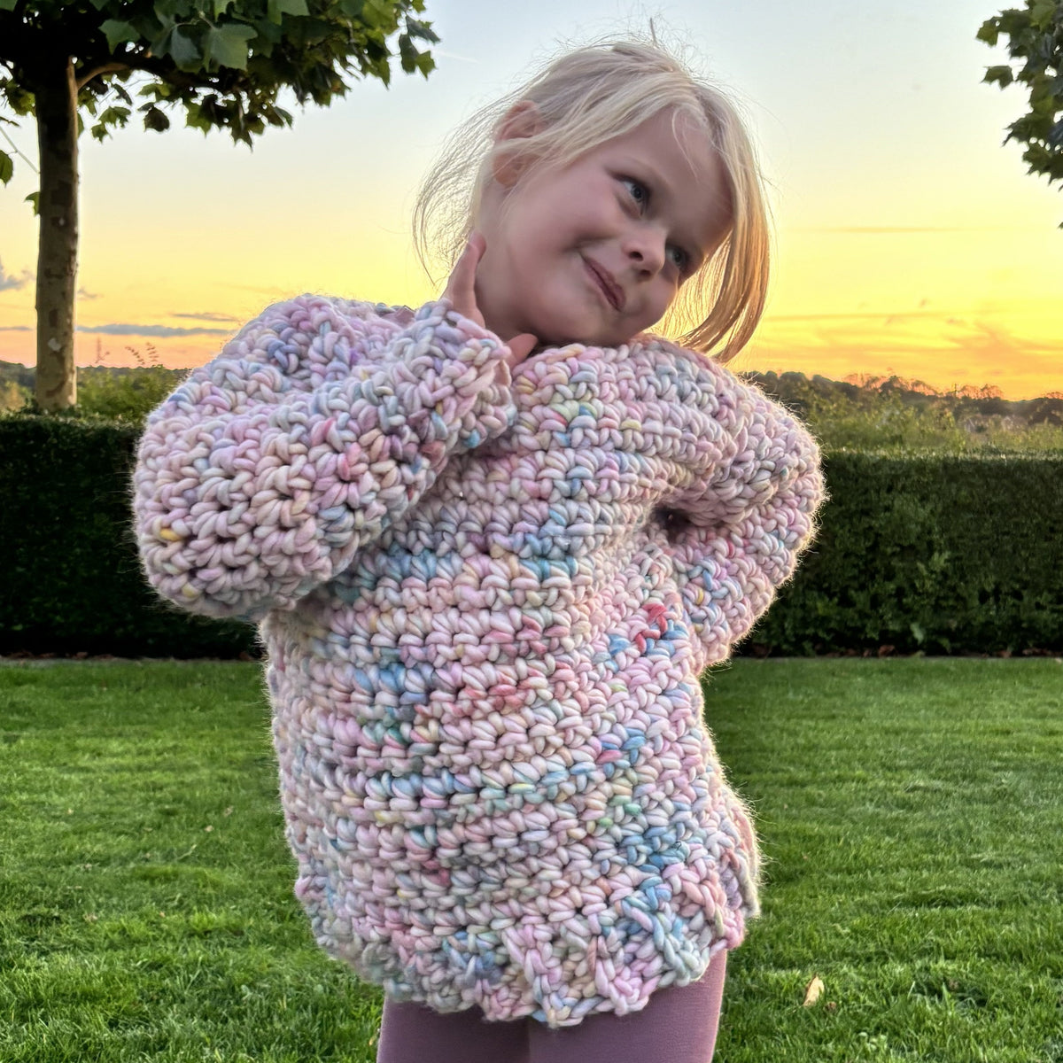 Smilende pige viser en hæklet børnesweater i lyserøde, lyseblå og lilla nuancer med chunky maskestruktur og ribkanter.