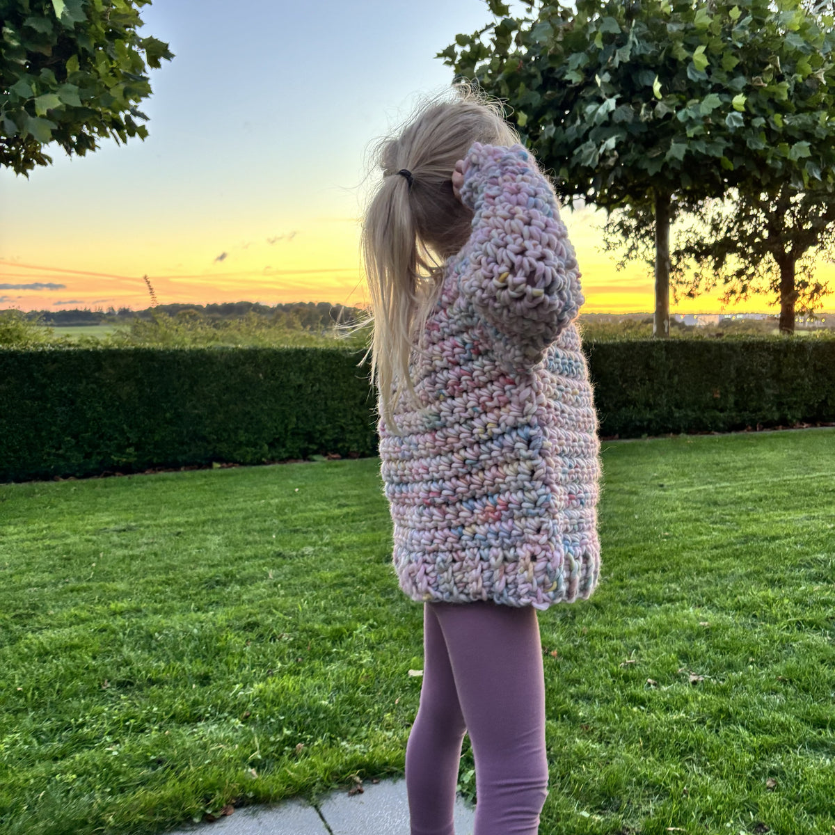 Hæklet Yarnicorn Sweater til børn set bagfra med pastelfarvet, tykt garn, lange ærmer og bred ribkant forneden.